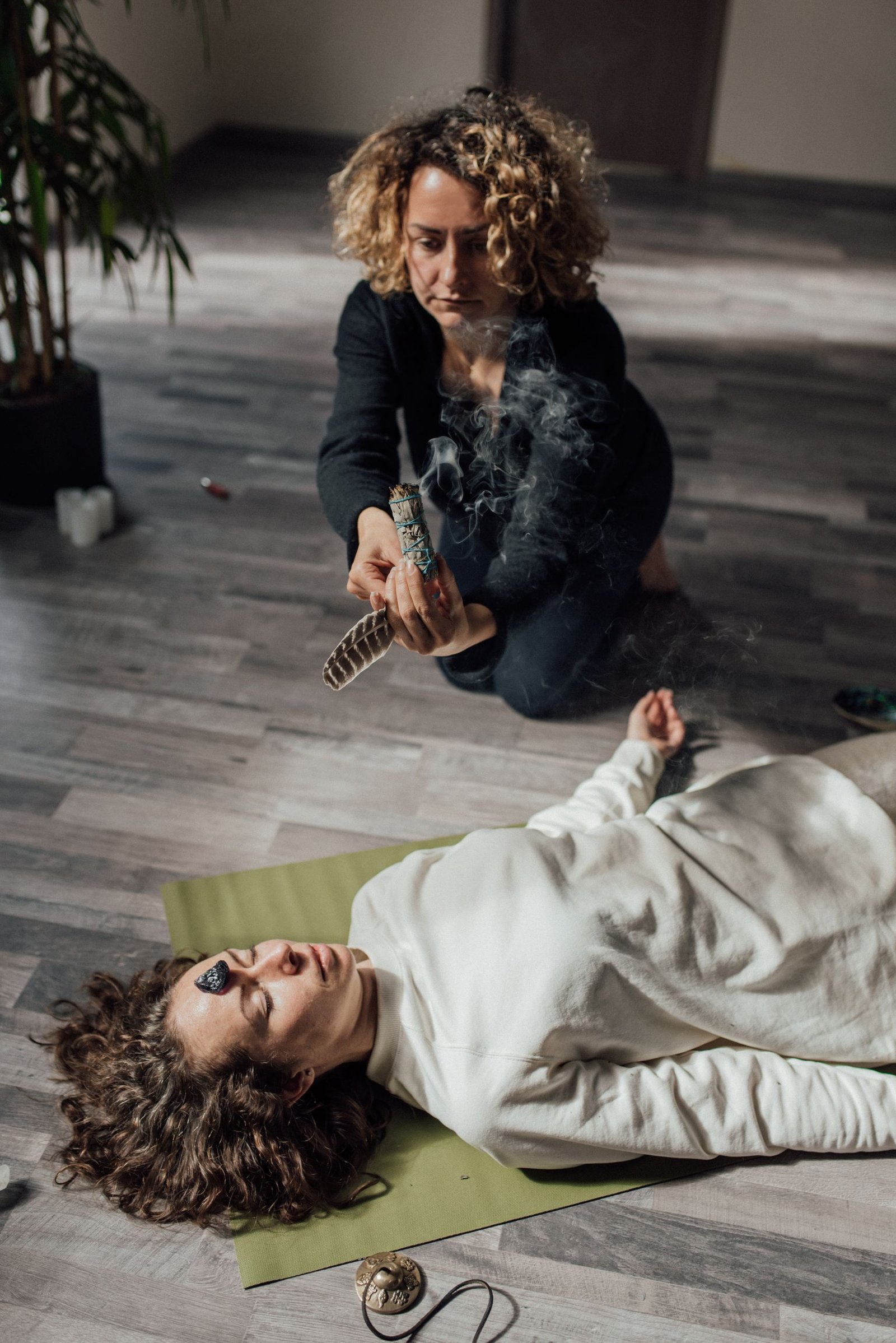 A woman performs a healing ritual with smudging and crystals on another woman indoors.